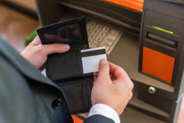 Önplanda bulanık ATM 'nin yanında dururken kredi kartını cüzdanına koyan işadamının kırpılmış görüntüsü