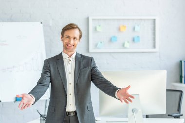 Front view of happy businessman with open arms looking at camera with blurred office on background clipart