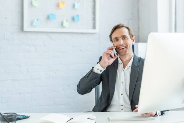 Gülümseyen işadamı kameraya bakıyor, telefonla konuşuyor, iş yerinde oturuyor.