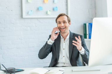 Neşeli iş adamı, iş yerinde telefonda konuşurken ön planda bulanık bilgisayar monitörü ile