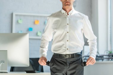 Cropped view of businessman showing empty pockets, while standing near workplace on blurred background clipart
