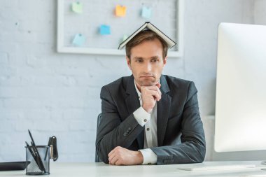 Upset businessman with notebook on head, sitting at workplace with blurred mesh organizer on background clipart