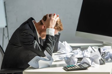 Stressed businessman with hands on head, sitting at workplace with crumbled papers on blurred background clipart