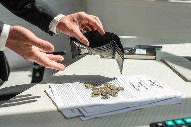 Cropped view of businessman holding empty wallet near coins on petitions for bankruptcy on table clipart