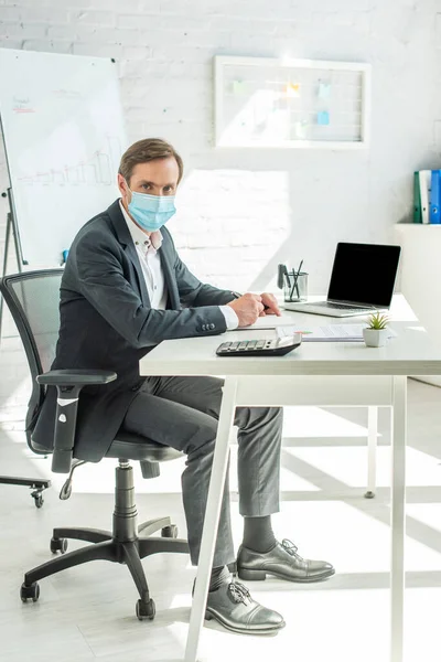 ノートを書きながら オフィスの職場に座っている間 医療用マスクのビジネスマンの完全な長さカメラを見て — ストック写真