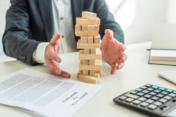 Cropped view of businessman with hands near blocks wood game, sitting at workplace on blurred background