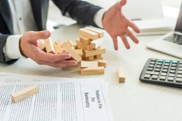 Cropped view of businessman with hands near block wood game falling near petition for bankruptcy on blurred background
