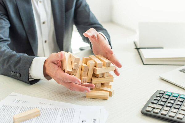 Cropped view of businessman holding blocks wood game collapsing, while sitting near documents at workplace 