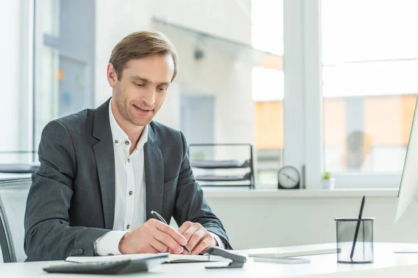 Χαμογελαστός Επιχειρηματίας Που Γράφει Στο Σημειωματάριο Ενώ Κάθεται Στο Τραπέζι — Φωτογραφία Αρχείου
