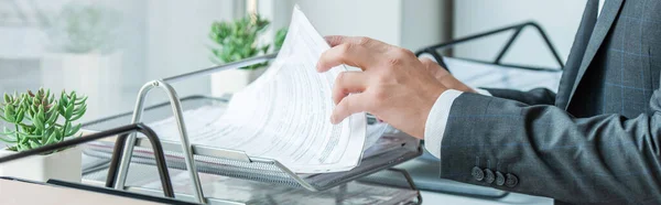 Cropped view of businessman looking for paper in document tray, while standing near windowsill, banner