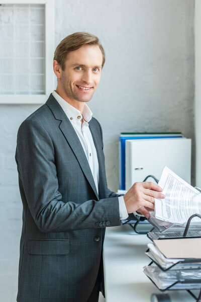 Happy businessman looking at camera, while searching paper in document tray on windowsill