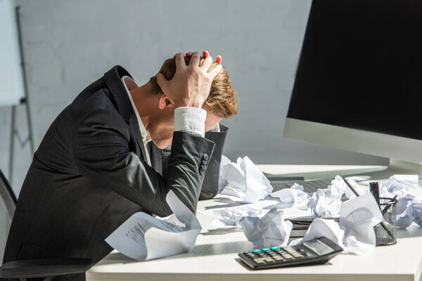 Стресс бизнесмен с руками на голове, сидя на рабочем месте с крошечными бумагами на размытом фоне