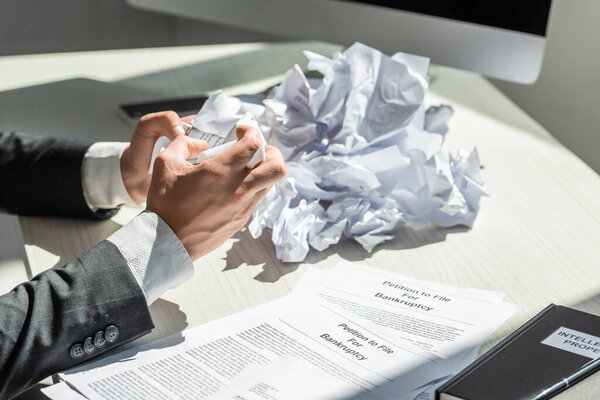 Cropped view of businessman holding crumbled paper near petitions for bankruptcy at workplace on blurred background
