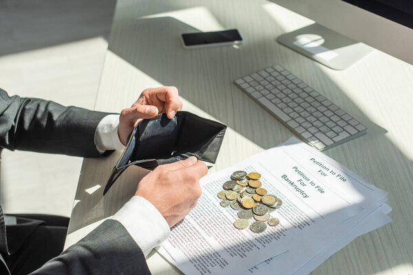 Cropped view of businessman holding empty wallet near coins and petitions for bankruptcy on workplace