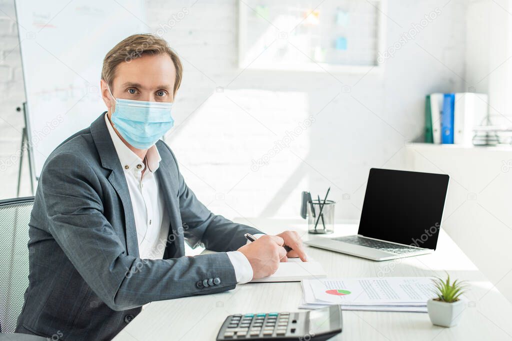 Businessman in medical mask looking at camera, while writing in notebook at workplace on blurred background