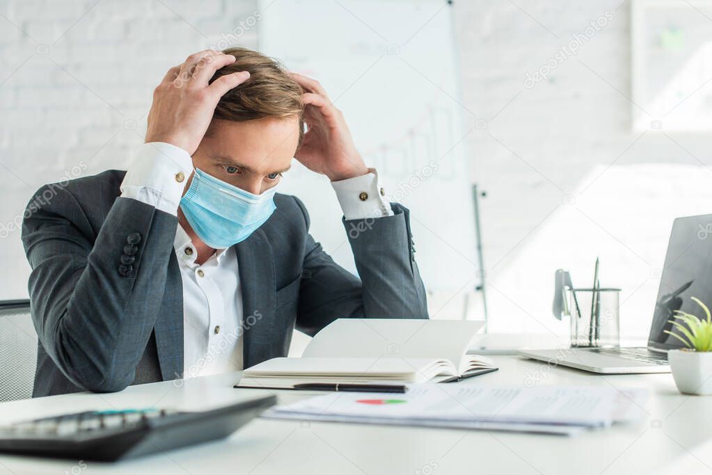 Bankrupt in medical mask holding hands near head, while sitting at workplace with blurred flipchart on background