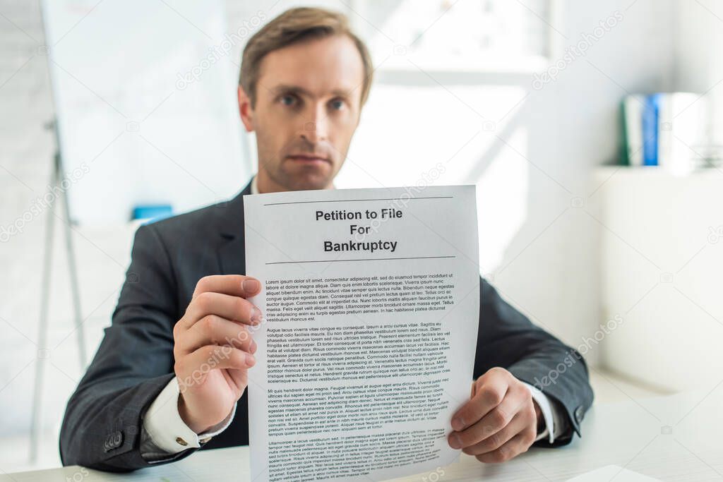 Serious businessman looking at camera and showing petition for bankruptcy, while sitting at desk on blurred background