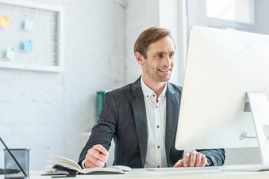 Happy businessman looking at computer monitor, while sitting at workplace in office on blurred background