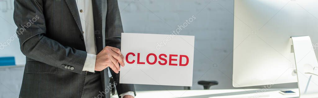 Cropped view of businessman holding sign with closed lettering, while standing near workplace on blurred background, banner