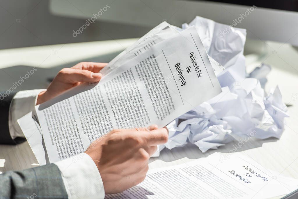 Cropped view of businessman holding petition for bankruptcy near pile of crumbled papers on blurred background