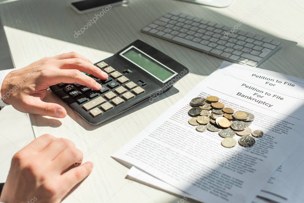 Cropped view of businessman counting on calculator near coins on petitions for bankruptcy on table