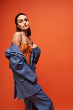 Esmer kadın, turuncu bir arka plana karşı modaya uygun mavi bir takım elbise ve korse sergiliyor..