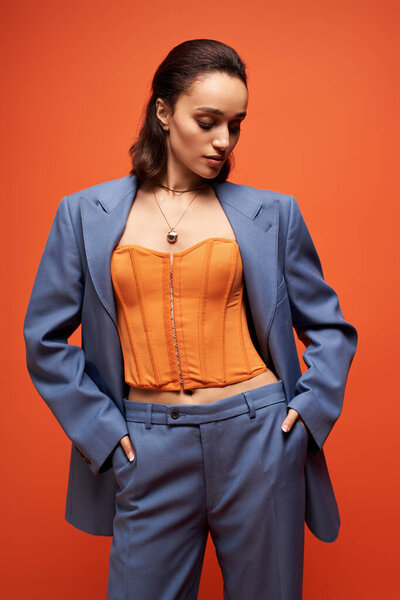 Молодая брюнетка уверенно позирует в модном синем костюме с оранжевым корсетом, излучая элегантность.