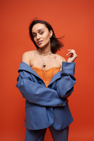 Брюнетка позирует в синем костюме с оранжевым корсетом на ярком фоне.