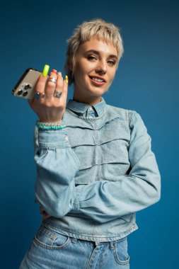 Şık, kısa saçlı genç bir kadın telefonunu modern bir ortamda tutarken kendine güvenerek gülümsüyor..