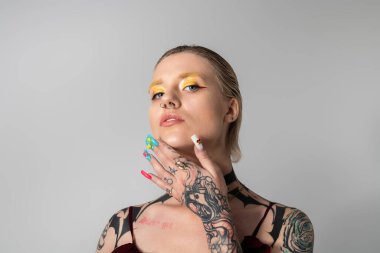 Kendinden emin genç bir kadın, tarafsız bir zemine karşı cesur makyaj ve renkli tırnaklar sergiliyor..