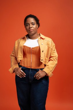 Kendine güvenen, iri yarı, şık bir sonbahar elbisesi giyen siyahi bir kadın canlı bir arkaplanda duruyor..