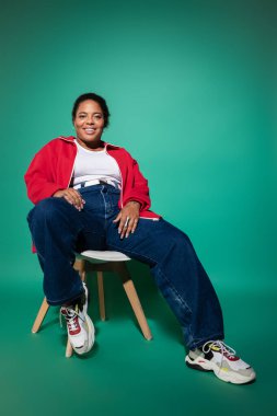 Kendinden emin, iri yarı, siyah bir kadın şık bir sonbahar elbisesi içinde canlı bir arka planda poz veriyor..