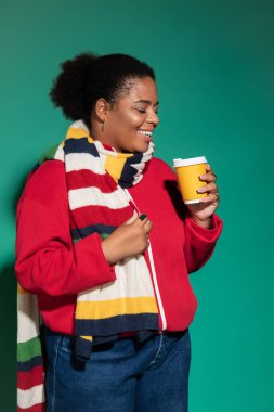 Parlak giyimli bir kadın stüdyo ortamında sıcak bir içecek tutarken neşe saçıyor..