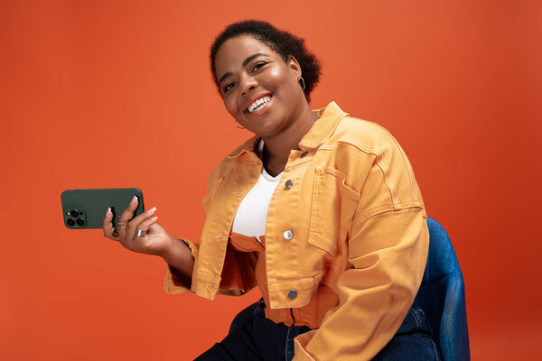 Bright orange backdrop highlights a fashionable plus size woman in a trendy autumn outfit, smiling.