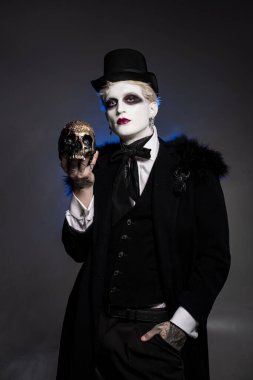 Yakışıklı genç bir adam zarif bir poz veriyor, büyüleyici bir Cadılar Bayramı kostümü giymiş, elinde bir kafatası tutuyor..