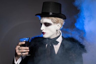 Özenle hazırlanmış Cadılar Bayramı kostümü içinde genç yakışıklı bir adam etrafı dumanla çevrili ürkütücü bir içki tutuyor..