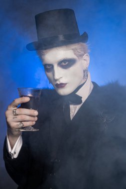 Dramatik bir Cadılar Bayramı kostümü giymiş genç ve yakışıklı bir adam dumanlı bir atmosferde bardak tutuyor..