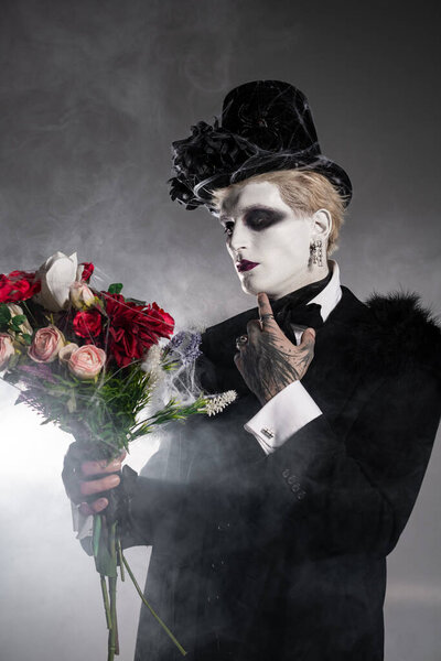 A young man in a creative Halloween costume holds a bouquet of roses against a smoky backdrop.