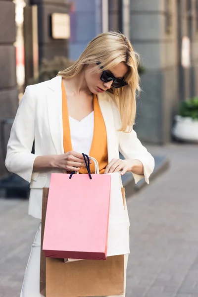 Stylish young woman in sunglasses looking into shopping bags on street — Stock Photo