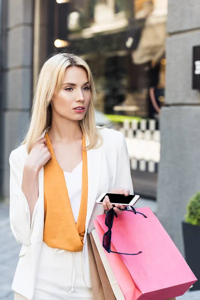 Attraktive blonde Frau mit Sonnenbrille, Smartphone und Einkaufstaschen, die auf der Straße wegschaut — Stockfoto