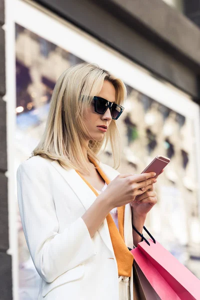 Bela menina loira elegante em óculos de sol segurando sacos de compras e usando smartphone — Fotografia de Stock