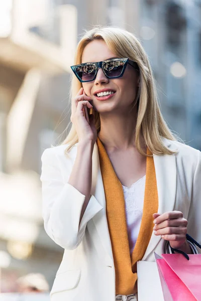 Schöne lächelnde junge Frau mit Sonnenbrille, Einkaufstüten in der Hand und im Gespräch mit dem Smartphone — Stockfoto