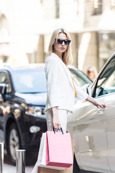 Стильна молода жінка в сонцезахисних окулярах тримає сумки і відкриває автомобіль — стокове фото