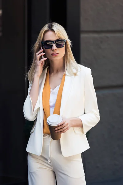 Stylish young woman holding disposable coffee cup and talking by smartphone on street — Stock Photo