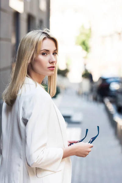 Belle jeune femme blonde élégante tenant des lunettes de soleil sur la rue — Photo de stock