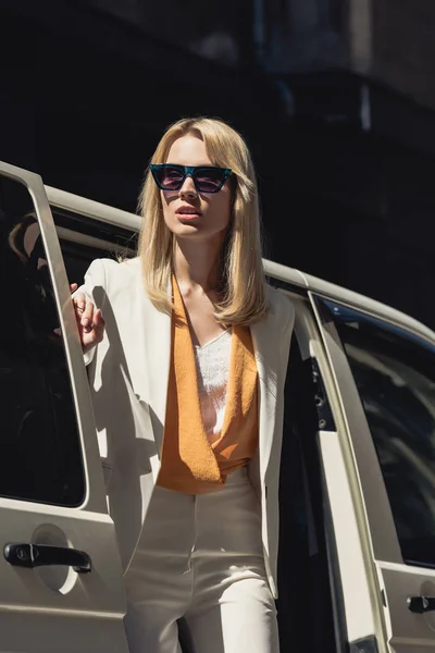Стильна молода жінка в сонцезахисних окулярах відкриває двері автомобіля і дивиться вбік — стокове фото