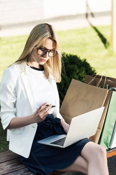 Young woman holding credit card and using laptop while sitting on bench with shopping bags — Stock Photo