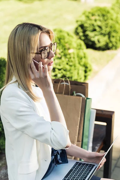 Seitenansicht eines stilvollen Mädchens, das mit Smartphone und Laptop spricht, während es mit Papiertüten auf einer Bank sitzt — Stockfoto