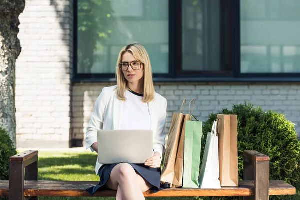 Young blonde woman in eyeglasses using laptop while sitting on bench with shopping bags — Stock Photo