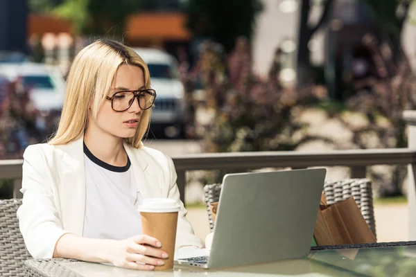 Молодая блондинка в очках с одноразовой чашкой кофе и ноутбуком — стоковое фото